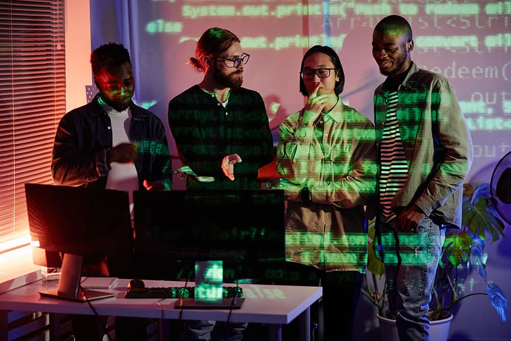 Group of four intercultural software engineers standing by desk with computer