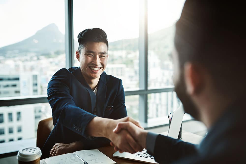 Shot of two young businessmen shaking hands while sitting at a desk in a modern office.